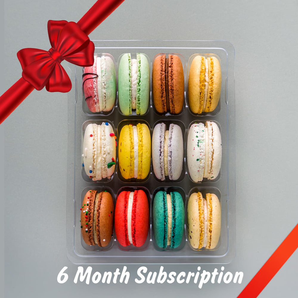 Assorted colorful macarons in a clear container with a red ribbon on a gray background, labeled '6 Month Subscription'.