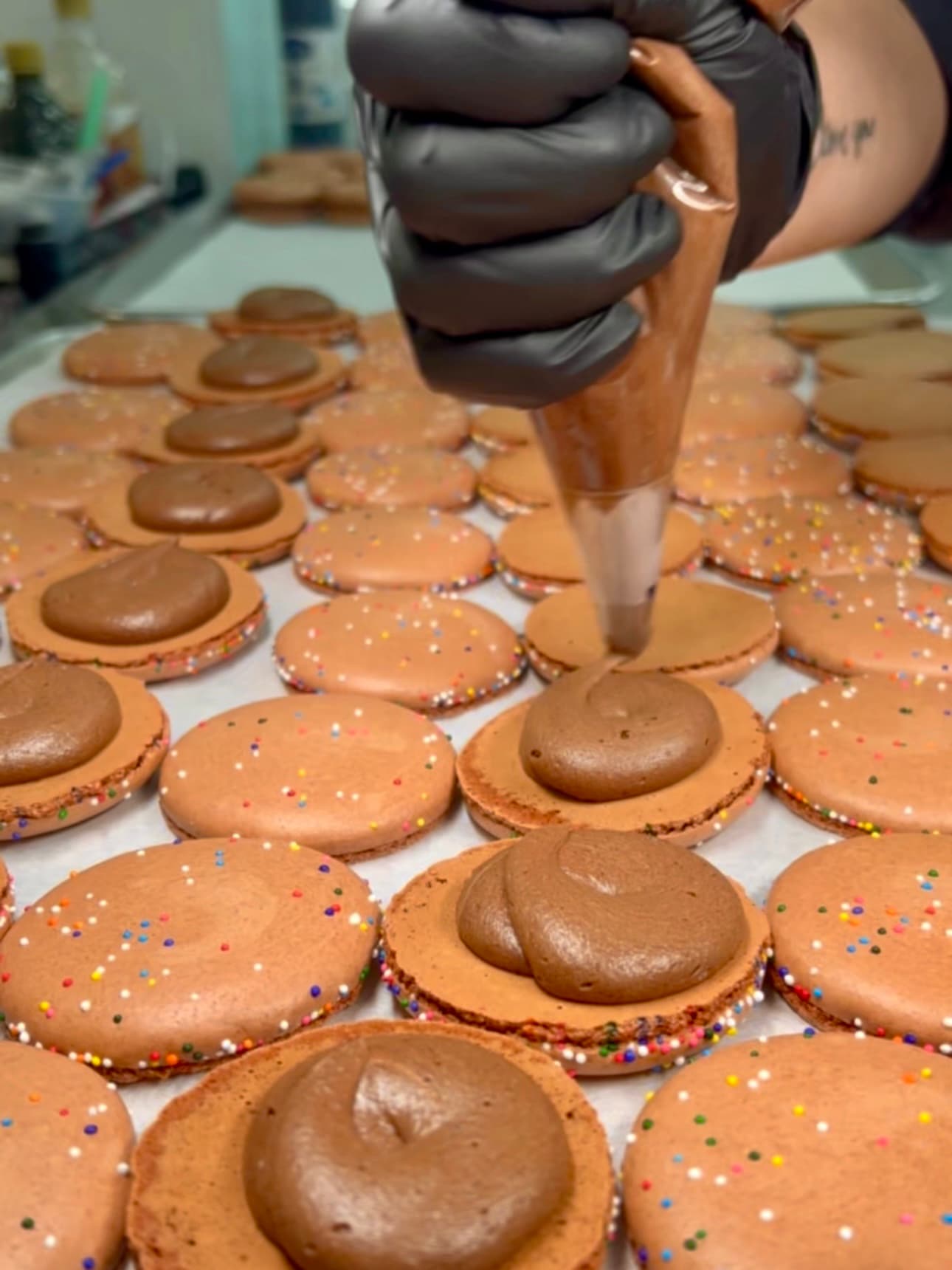 Piping chocolate buttercream onto brown macaron shells with rainbow nonpareil sprinkles