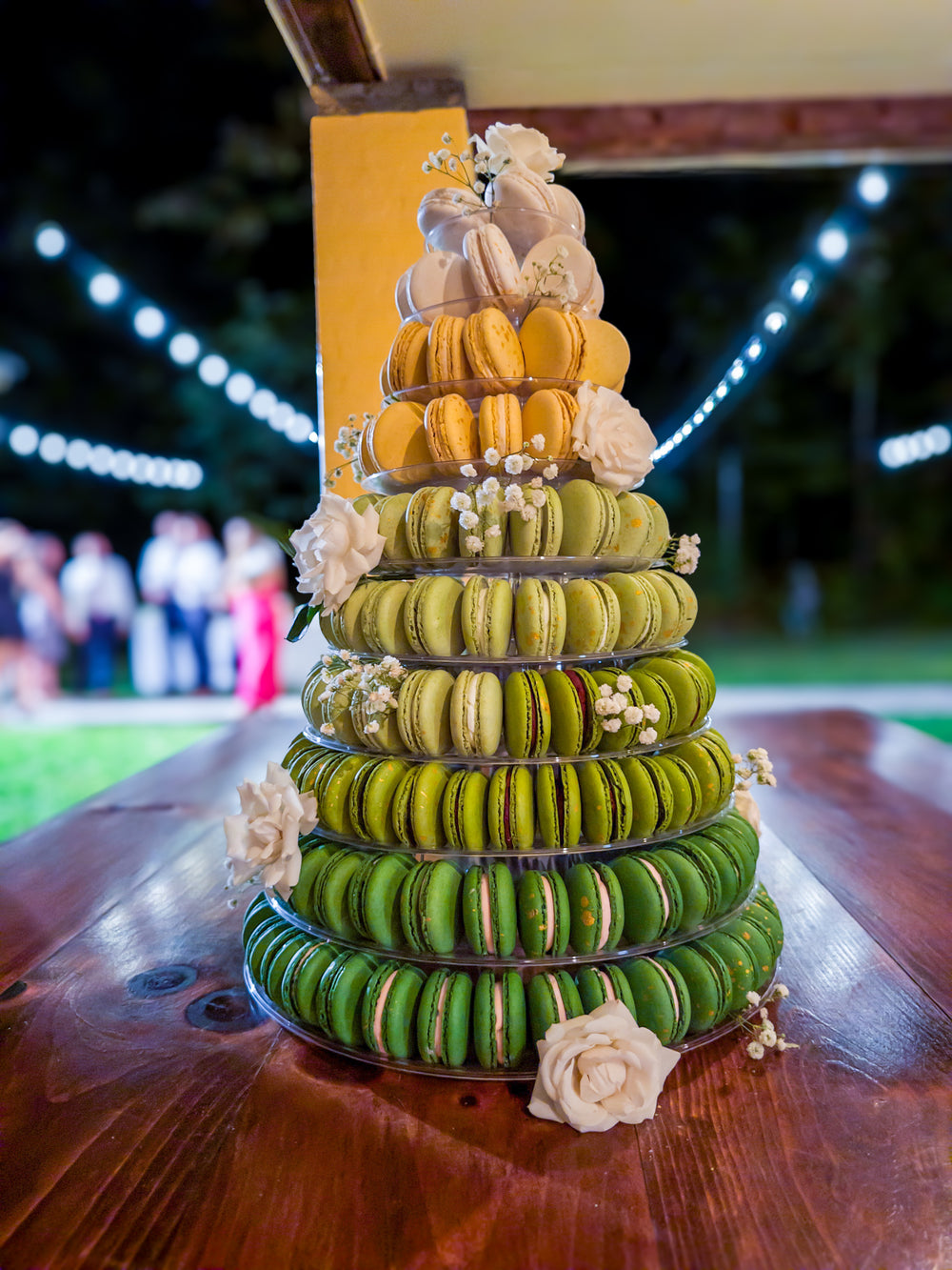 Multilevel cake made of green, yellow, and orange macarons on a wooden table with blurred lights in the background.