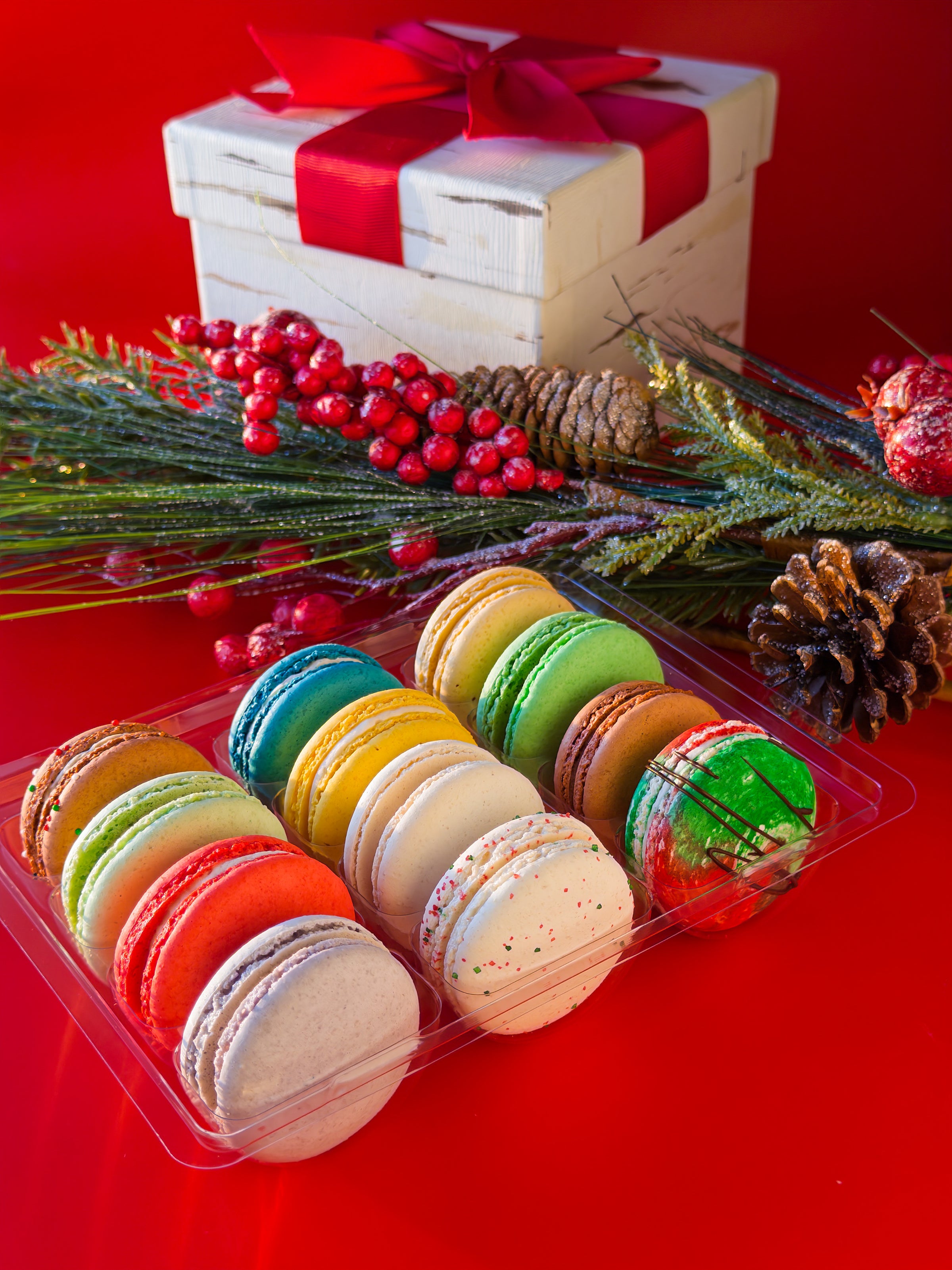 Colorful macarons in a clear container with a Christmas-themed gift box and decorations on a red background.