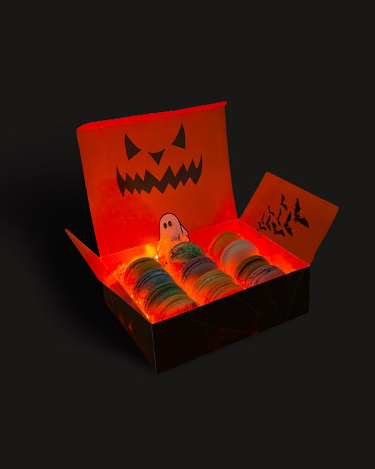 A 12 pack of French macarons inside a glowing orange box with a jack-o’-lantern face on the box