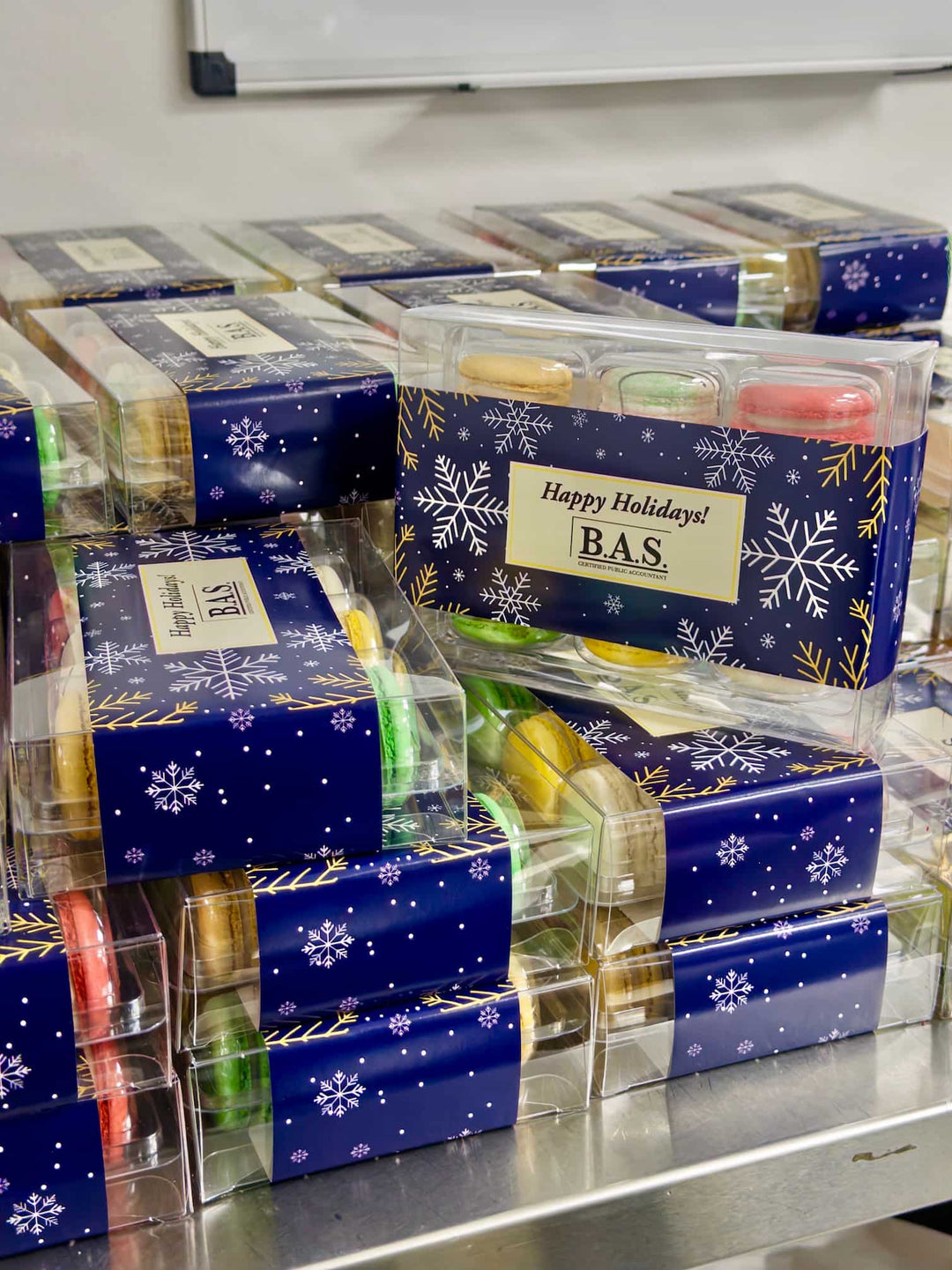 Packaged macarons in blue and clear boxes with 'Happy Holidays' and 'BAS' labels.